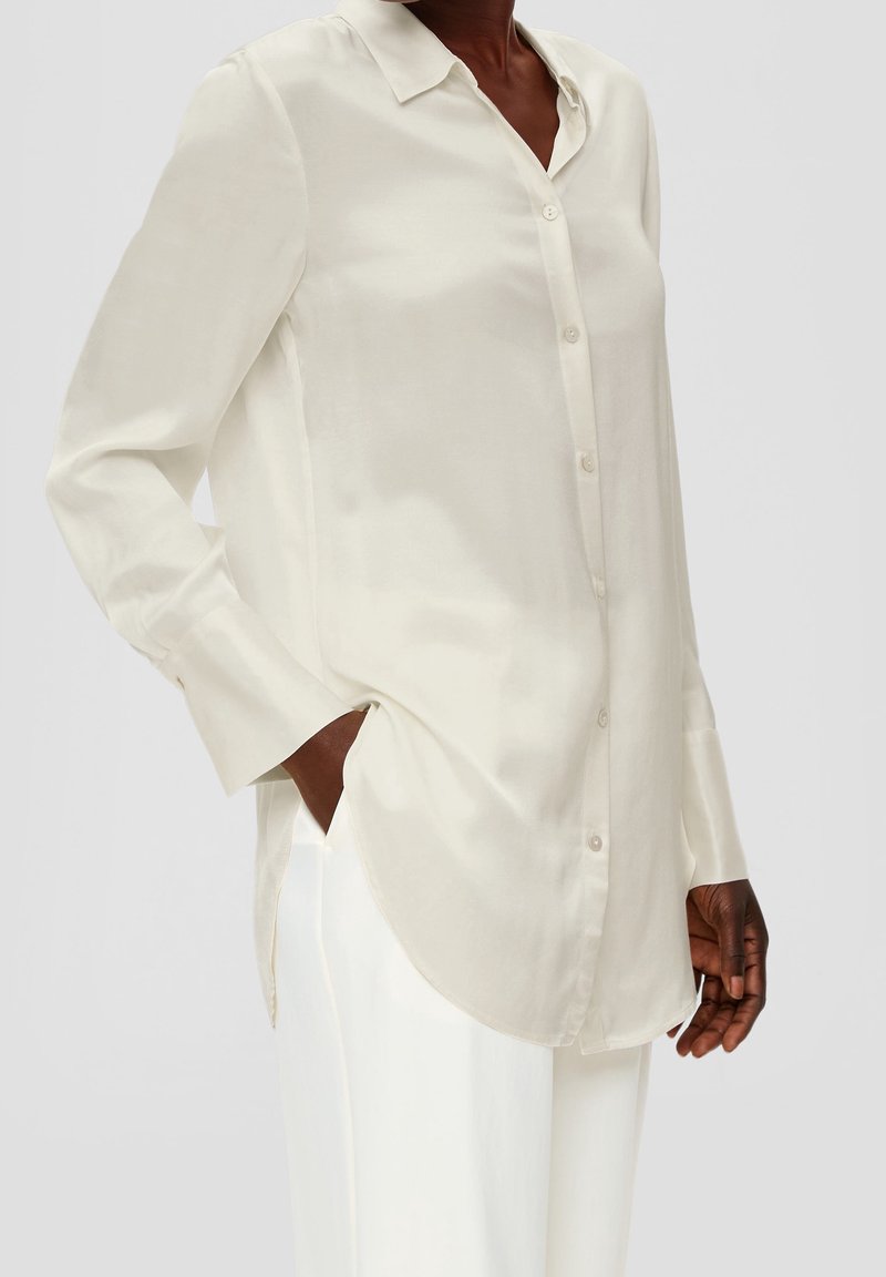 Femme portant une chemise écrue à manches longues boutonnée, main dans la poche, associée à un pantalon blanc sur un fond uni.