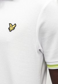 Fehér, texturált anyagból készült póló, melyen sárga hímzett madárlogó található a mellrészen. A ujjain zöld-fehér csíkos díszítések.