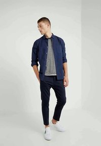 Chemise bleu marine boutonnée par-dessus une chemise rayée, associée à un pantalon slim-fit bleu marin et des baskets blanches. Design simple, texture du tissu non visible.