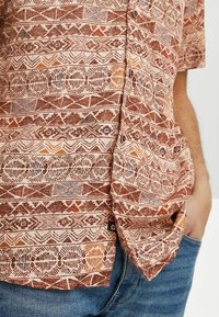 Camisa de manga corta hecha de tejido texturizado que presenta un patrón tribal repetitivo en tonos tierra de marrón, naranja y azul. Con botones en la parte delantera.