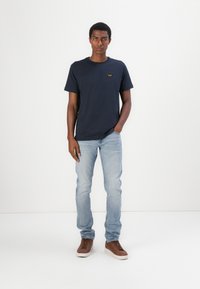 Navy blue cotton t-shirt with a round neckline and small chest logo, paired with light blue jeans and brown shoes in a standing pose.