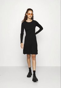 Melns garu piedurkņu kleita ar pieguļošu augšdaļu un izplešanos apakšā. Modelis valkā melnas zeķes un masīvas melnas kurpes, kā arī oranžus saulesbrilles.