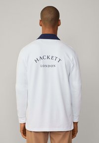 Valkoinen pitkähihainen polo, jossa on tummansininen kaulus. "HACKETT LONDON" painettuna tummansinisellä selkäpuolella. Pehmeä puuvillamateriaali, rento malli.