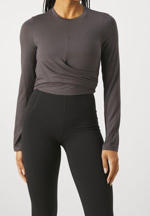Vrouw draagt een aansluitend donkergrijs topje met lange mouwen en een gedraaide detail en zwarte leggings met hoge taille, staand tegen een effen achtergrond.