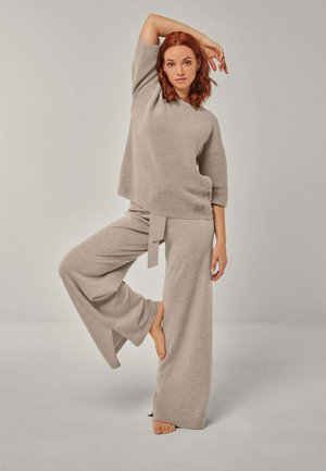 Woman with red hair wearing a beige knit top and wide-leg pants, balancing on one foot with one arm raised above her head.