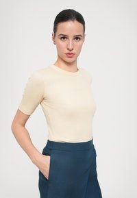 Upravené tričko s krátkým rukávem a ribovaným vzorem v světlé béžové barvě, které je kombinováno s kalhotami s vysokým pasem v tmavě modré barvě s hladkým povrchem a bočními kapsami.