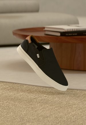 Czarny sneaker typu slip-on z białą podeszwą i brązową zawieszką na pięcie, umieszczony obok drewnianego stolika kawowego ze stosami książek na jasnym dywanie i w tle sofa.