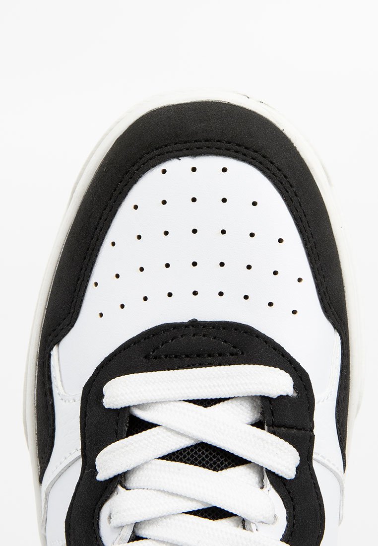 Gros plan sur l'embout d'une sneaker noir et blanc avec du cuir blanc perforé et des lacets blancs sur un fond blanc.