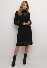 Rochie neagră din dantelă, cu mâneci lungi și puffate, guler înalt și detalii cu nasturi. Materialul are un model texturat și un tiv până la genunchi.