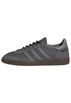 Niskoprofilowy szary sneaker Adidas z panelami z zamszu i siateczki, trzema paskami po bokach, brązową fakturowaną podeszwą i metką z tkaniny Cordura na pięcie.