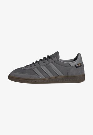 Zapatilla Adidas gris de perfil bajo con paneles de ante y malla, tres rayas en el lateral, suela marrón texturizada y etiqueta de tela Cordura en el talón.