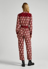 Jumpsuit rouge à motifs avec manches longues, taille ceinturée et col contrasté. Présente un motif géométrique complexe avec des accents crème.