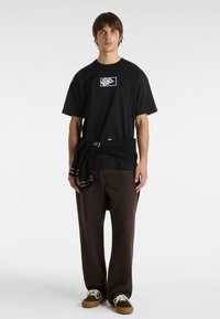 Camiseta de algodón negra con un logo blanco en el pecho, combinada con pantalones anchos marrones y zapatillas marrones. Suéter atado a la cintura.