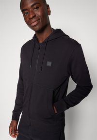 Svart zip-up hoodie med huva, tillverkad av texturerat tyg. Har en framficka och en liten logotyp patch på bröstet.