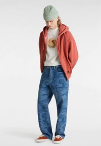 Rode hoodie met rits, wit T-shirt met cirkelvormig ontwerp, blauwe camo-spijkerbroek en oranje sneakers; groene beanie en oorbellen maken de look compleet.