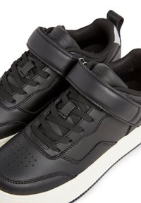 Paire de baskets en cuir noir avec lacets noirs, fermetures à scratch et semelles en caoutchouc blanches, présentées sur fond blanc.