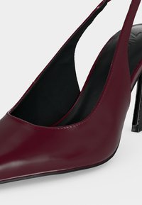Escarpin slingback en cuir bordeaux à talon haut avec bout pointu et semelle intérieure noire sur fond clair.