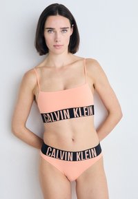Calvin Klein bikini-set in zachte perzik, met een vierkante crop top met zwarte logoband en bijpassende hoge taille broekjes met vergelijkbare branding.