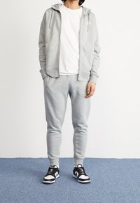 Person klädd i en ljusgrå Nike träningsoverall med zip-up huvtröja, vit t-shirt och svarta och vita Nike sneakers står på en blå golv.