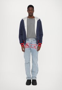 Mørkeblå jakke med røde detaljer, grå t-shirt, lyseblå jeans med en rød broderet design og sorte slip-on sko.