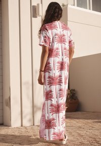 Lange witte jurk met een rood palmbomenpatroon, korte mouwen, losse pasvorm en zijsplitten, afgebeeld tegen een lichtgekleurde achtergrond.