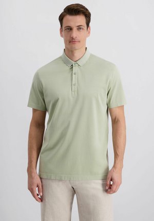 Mann trägt hellgrünes Polohemd mit kurzen Ärmeln und beigen Hosen, steht vor einfachem hellem Hintergrund und schaut nach vorne.