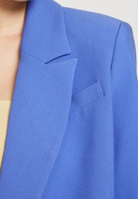 Blazer bleu avec un design structuré, des revers crantés et une poche poitrine unique. Le tissu présente une apparence lisse et texturée.