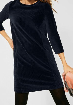 Kobieta ubrana w granatową sukienkę sztruksową z rękawami do połowy przedramienia, trzymająca w prawej ręce różowo-szary szalik w wzory.