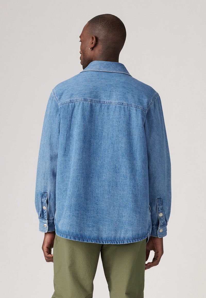 Chaqueta de mezclilla azul claro con cuello y puños abotonados, que presenta un ajuste holgado y detalle en el yugo trasero. Textura suave, dobladillo sin acabar.