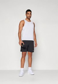 Bela športna majica brez rokavov, črne kratke hlače in beli supergi. V roki drži črno steklenico za vodo. Enostavna zasnova s kontrastnimi barvami.