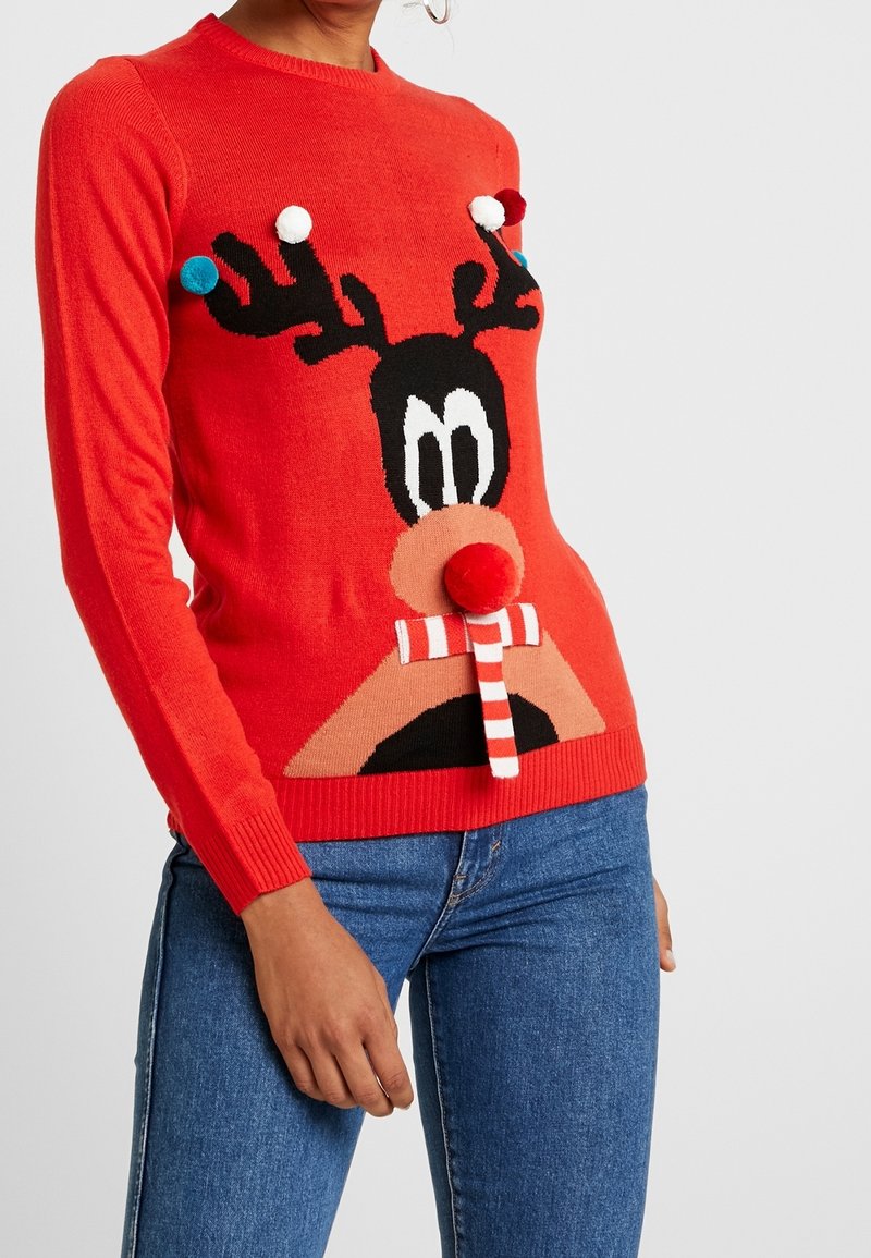 Pull rouge avec visage de renne en dessin animé, écharpe rayée, nez en pompon et pompons colorés sur les bois, porté avec un jean bleu.