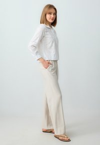 Witte blousetop met lange mouwen, gedetailleerde patronen en kraag, gecombineerd met lichtbeige wijde broekspijpen en bruine slippers.