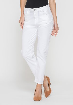 Donna che indossa pantaloni bianchi slim e scarpe marroni con tacchi alti, in piedi con una gamba leggermente incrociata davanti all'altra.