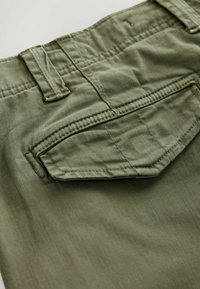 Gros plan sur un pantalon en tissu vert montrant une poche arrière avec rabat et une boucle de ceinture au niveau de la taille.