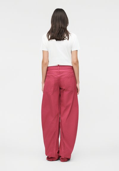 Γυναίκα με μακριά καστανά μαλλιά, που φοράει λευκό κοντομάνικο τοπ και φαρδιά κόκκινα παντελόνια, στέκεται γυρισμένη πλάτη σε απλό φόντο.