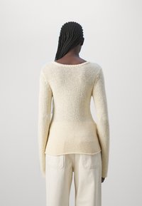 Pull en tricot crème avec un col large, silhouette ajustée, manches longues et motif texturé, associé à un pantalon de couleur claire.