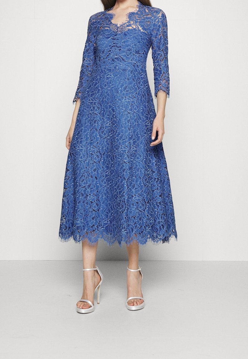 Robe en dentelle bleue avec manches trois-quarts, motif floral et ourlet festonné. Associée à des sandales à talons hauts argentées avec des lanières.