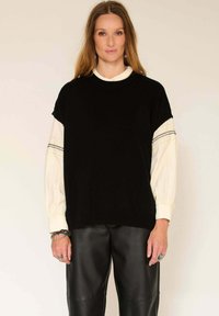 Pull noir à manches courtes superposé sur une chemise blanche avec des poignets côtelés. Porté avec un pantalon en cuir noir. Accessoire en argent.