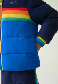 Kék puffertakó, amely multicolor vízszintes csíkokkal rendelkezik piros, narancs, sárga, zöld és kék színekben. Cipzáras záródással és bordázott mandzsettákkal rendelkezik.