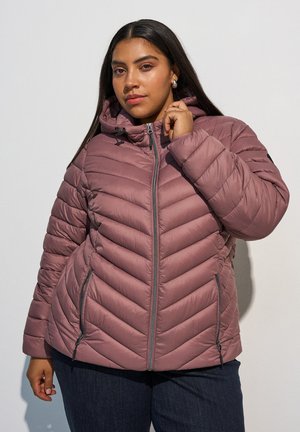 Vrouw met lang donker haar die een mauve doorgestikte capuchonjas en donkere broek draagt tegen een effen lichte achtergrond.