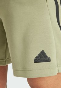 Olivově zelené šortky vyrobené z měkké, texturované tkaniny. Na spodní části vlevo se nachází černá logo nášivka Adidas. Rovný lem a detaily bočních švů.