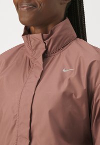 Giacca impermeabile rosa chiaro con colletto alto e chiusura a zip. Presenta un piccolo logo Nike swoosh argento sul lato sinistro.