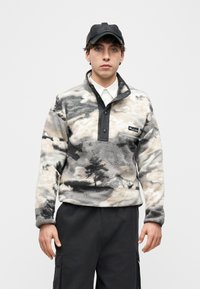 Νεαρός άνδρας που φοράει μαύρο καπέλο, λευκό πουκάμισο με γιακά, φλις μπουφάν σε γκρι και μπεζ μοτίβα και μαύρο παντελόνι cargo, με φόντο έναν απλό τοίχο.