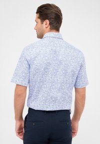 Korte mouwen shirt in lichtblauwe stof met een bloemenpatroon, voorzien van een normale kraag en een rugdetail. Gecombineerd met donkere broeken.