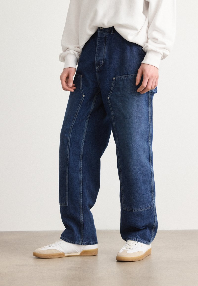 Jean large en denim bleu avec des poches frontales prononcées et des détails de couture visibles, associés à des baskets blanches sur un sol beige.