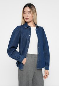 Camisa de denim com abertura frontal com botões, mangas longas bufantes e colarinho. Tecido azul-escuro com uma textura suave e botões prateados.