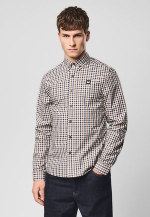 Junger Mann in einem langärmligen, karierten Hemd mit braunen, blauen und weißen Quadraten, der mit einer Hand in der Tasche seiner dunklen Jeans steht.