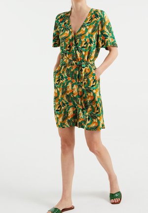 Femme portant une robe à manches courtes et col en V, avec des motifs de feuilles vertes et orange, dotée de poches latérales et d'une ceinture à la taille, associée à des sandales vertes à enfiler.