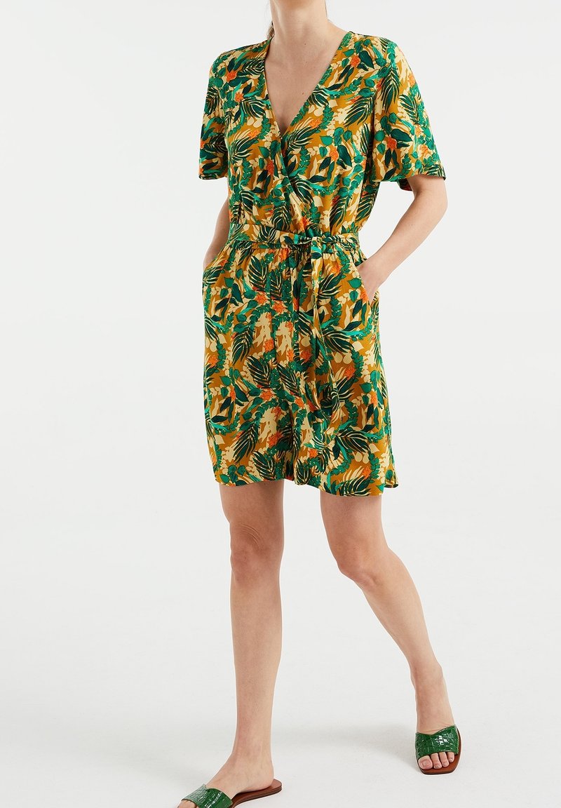 Femme portant une robe à manches courtes et col en V, avec des motifs de feuilles vertes et orange, dotée de poches latérales et d'une ceinture à la taille, associée à des sandales vertes à enfiler.