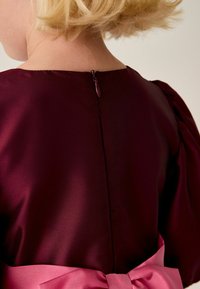 Burgunderfarbenes Satin-Kleid mit Puffärmeln, ausgestattet mit einem Reißverschluss hinten und einem breiten rosa Satinschleifenband, das in der Taille gebunden ist. Glatte Textur.